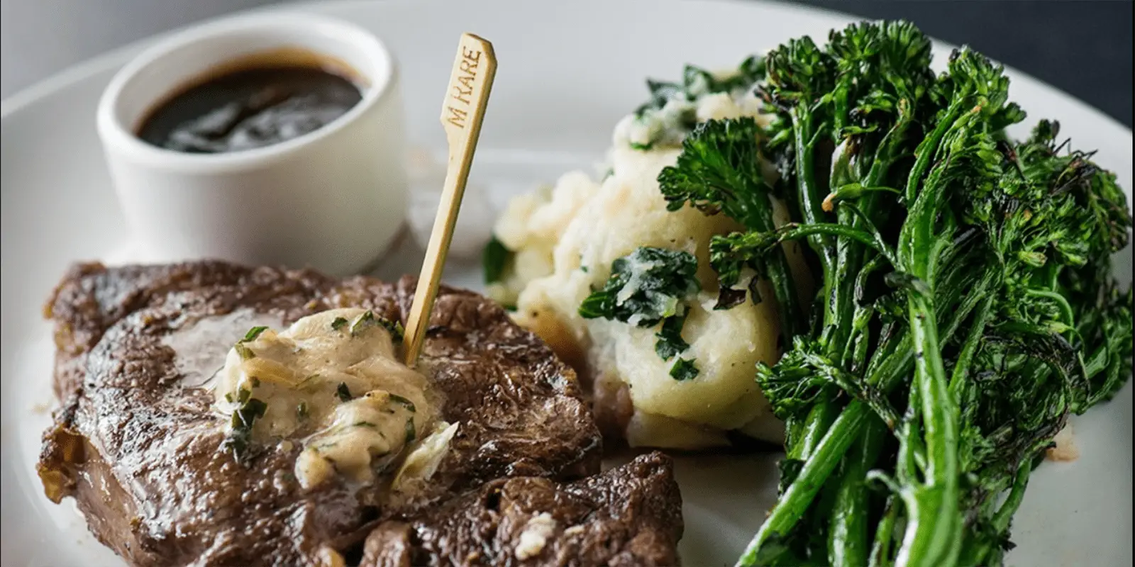 Plate of grilled steak topped with herb butter, a stick labeled "medium rare" inserted in the steak. Served with a side of cauliflower and broccolini. A small cup of dipping sauce sits in the background, making this dish a highlight of the best gluten-free dining in Waikiki.