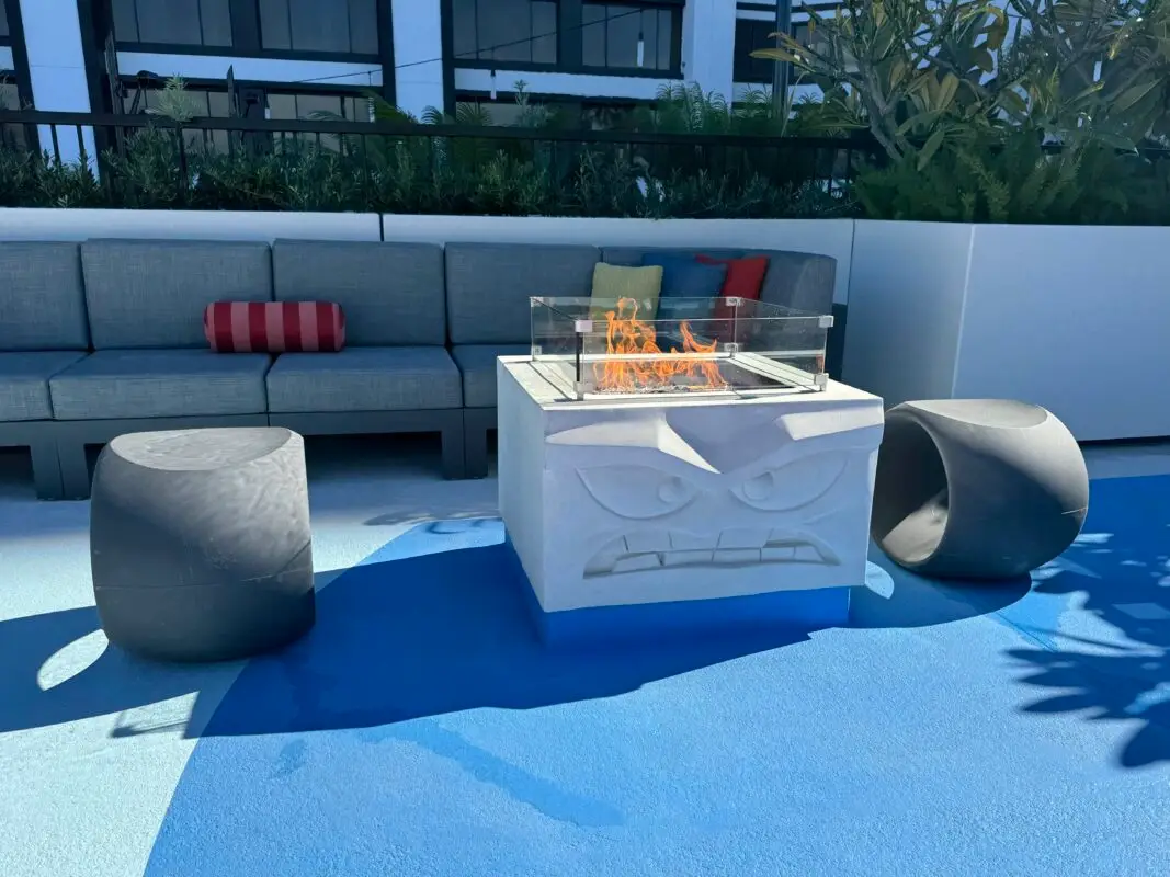 A modern outdoor patio features a square fire pit with glass panels and a carved angry face design, perfect for Pixar Fest gatherings. Surrounding the fire pit are two black cylindrical stools and a grey sectional couch adorned with colorful pillows. The ground is painted in vibrant shades of blue.