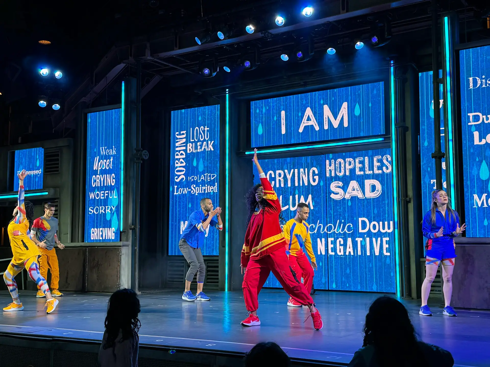 Performers on a brightly lit stage dance energetically. The background features large screens displaying words like "Crying," "Hopeless," "Sad," and "Negative." Dressed in colorful outfits, the setting suggests an emotional or motivational theme, reminiscent of the vibrant atmosphere at Pixar Fest.