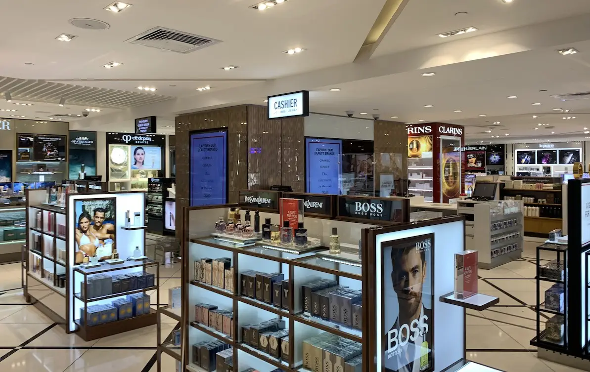A well-lit Duty Free Shops Hawaii retail store interior, displaying various shelves and counters filled with different brands of perfumes, skincare, and cosmetic products. A sign reading "CASHIER" is visible at the center of the store. Shiny tiled floor and modern décor.