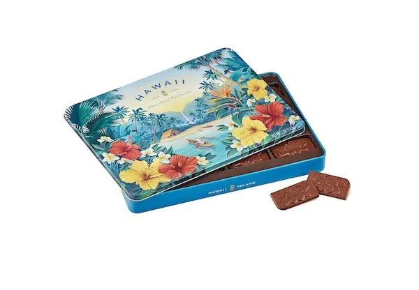 A decorative tin box labeled "Hawaii," often found in Duty Free Shops, features a tropical beach scene with palm trees, mountains, and hibiscus flowers. The lid is partially open, revealing several rectangular pieces of chocolate with a floral design embossed on them inside.