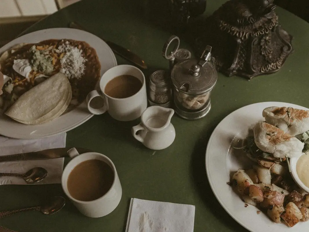 A dining table set with two plates of the best breakfast in Big Sur, featuring a dish with a tortilla and beans, and another with potatoes and a sandwich or bagel. The table also has two cups of coffee, a small jug, salt and pepper shakers, a sugar container, and cutlery on napkins.
