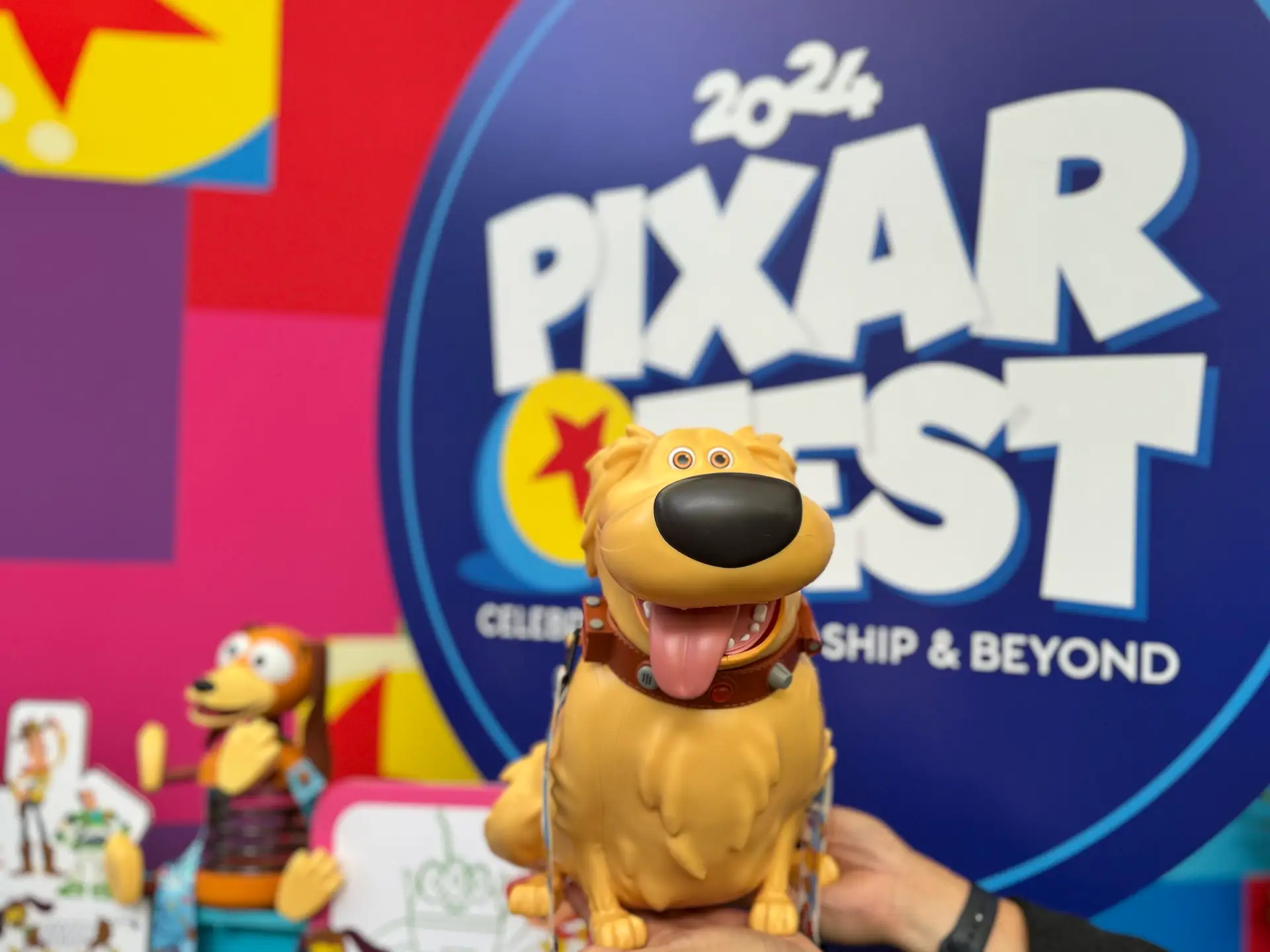 A hand holds a toy figure of Dug, the dog from Pixar's "Up," against a colorful backdrop featuring the "Pixar Fest 2024" logo. The background includes vibrant patterns and another toy resembling Woody from "Toy Story," highlighting the festivities at Pixar Fest.
