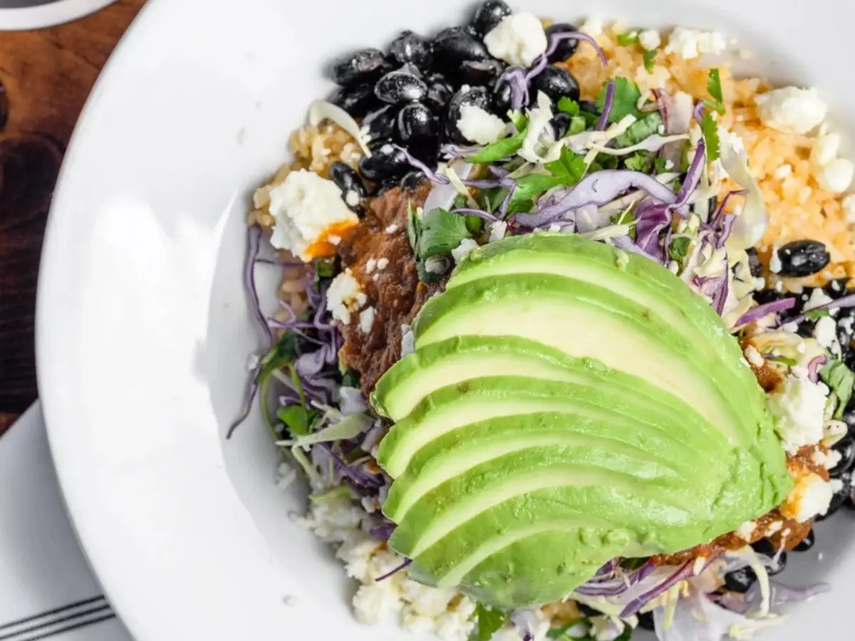 A bowl filled with a variety of fresh ingredients including a large fan of sliced avocado on top, black beans, crumbled feta cheese, thinly sliced purple cabbage, cilantro, and rice. This vibrant dish is not only colorful but also hailed as the Best Breakfast in Santa Cruz—truly a fresh, healthy meal option.