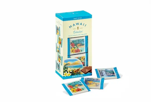 A box of Hawaii Collection milk chocolate squares is shown, available exclusively in Duty Free Shops Hawaii. The light blue and yellow packaging features tropical imagery. Three unwrapped chocolate squares with colorful wrappers depicting Hawaiian scenes are displayed beside the box.