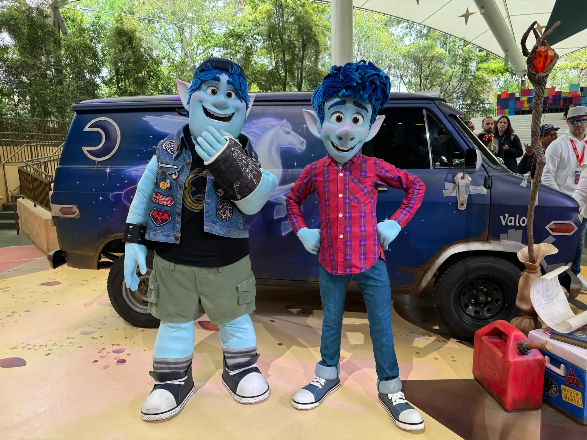 Two people dressed as blue-skinned characters from an animated film stand in front of a themed van at Pixar Fest. One wears a sleeveless vest with shorts, while the other sports a red plaid shirt with jeans. The van, adorned with a unicorn painting, is set against the backdrop of lush trees.