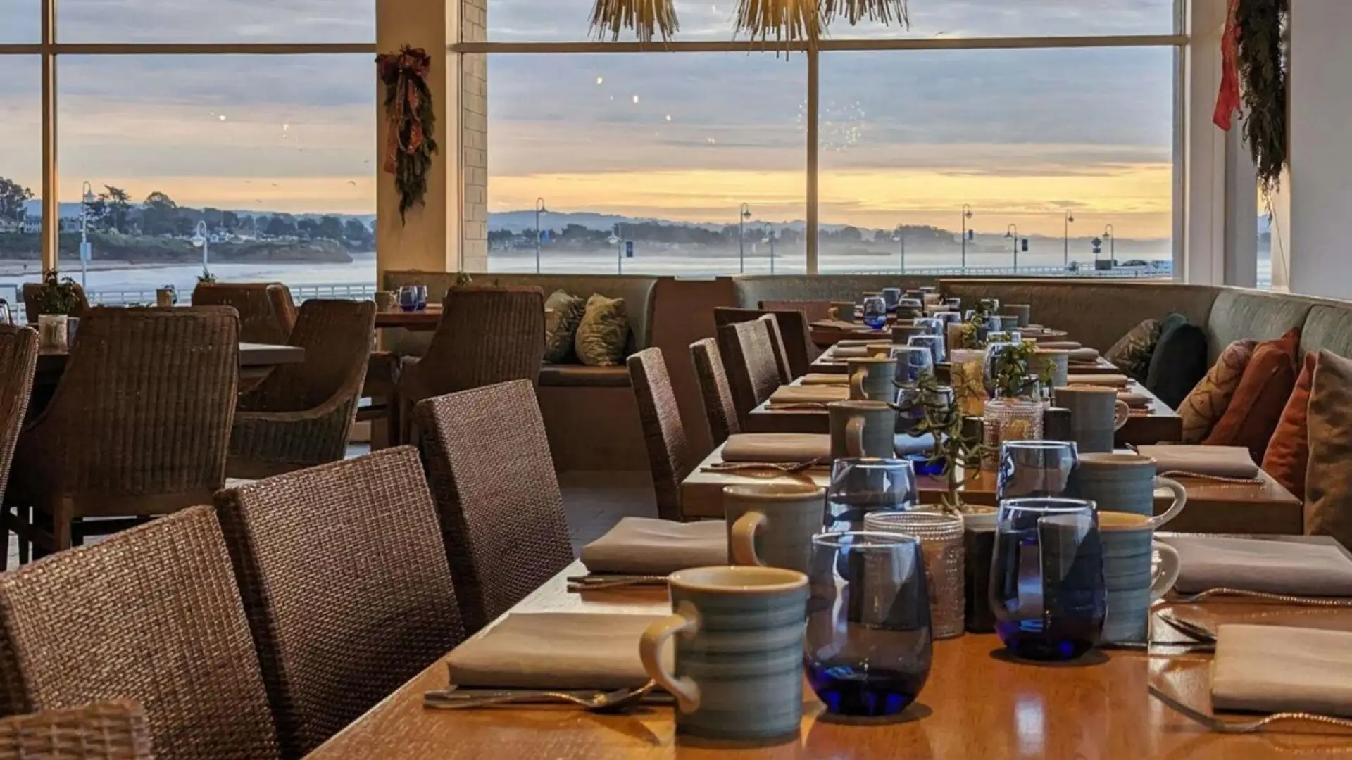 A waterfront restaurant's dining area with wooden tables set with blue-tinted glasses and ceramic mugs overlooks the calm sea at sunset. The spacious setup at Ocean View Santa Cruz features wicker chairs and expansive windows framing the scenic view.