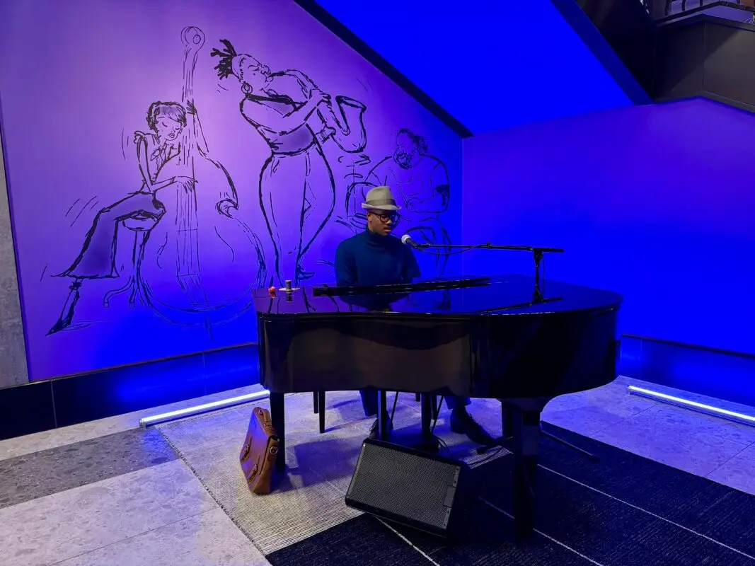 A person wearing a hat and glasses is playing a grand piano in a blue-lit room. Behind them, there is a mural of jazz musicians, including a bassist, a saxophonist, and a drummer, evoking the spirit of Pixar Fest. A small bag rests on the floor beside the piano.