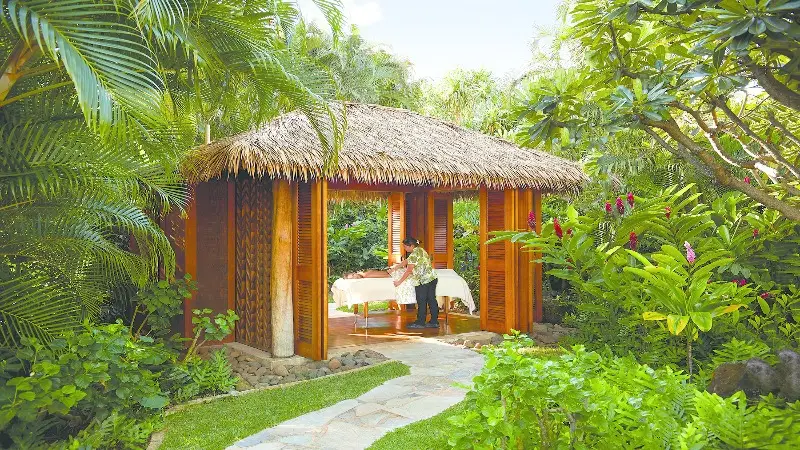 A person is preparing a table inside an open wooden hut with a thatched roof, surrounded by lush green tropical plants and flowers. A stone path leads to the hut, creating a serene and picturesque setting reminiscent of the best spa hotels Kauai has to offer.