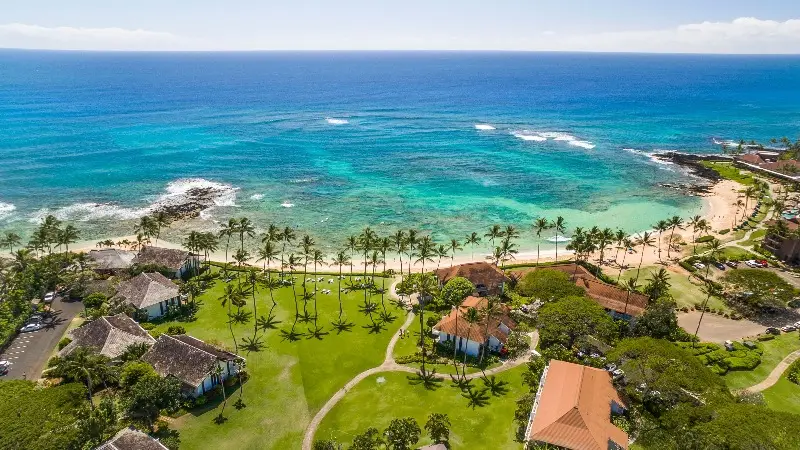 Aerial view of a coastal area with turquoise waters, waves breaking near the shore, a sandy beach lined with palm trees, and several buildings with red and white roofs surrounded by lush green lawns and pathways—truly one of the best oceanfront hotels Kauai has to offer.