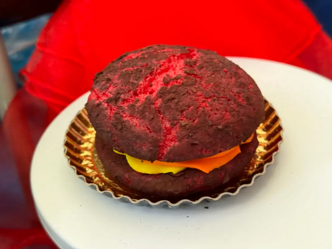 A round chocolate cookie sandwich filled with bright yellow cream, set on a decorative gold scalloped paper on a white surface. The cookie has a red tint, suggesting a mix of red and chocolate coloring, reminiscent of the playful hues seen at Pixar Fest.