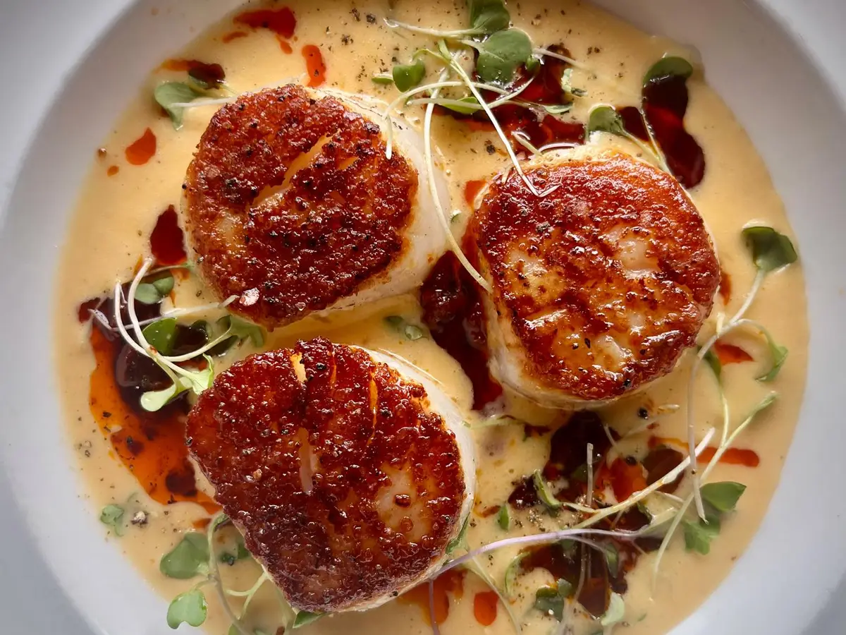 Three seared scallops are elegantly presented on a plate, surrounded by a creamy sauce and garnished with microgreens. Red oil droplets are drizzled artistically around the dish, adding color and contrast to this exquisite Seafood Santa Cruz creation.