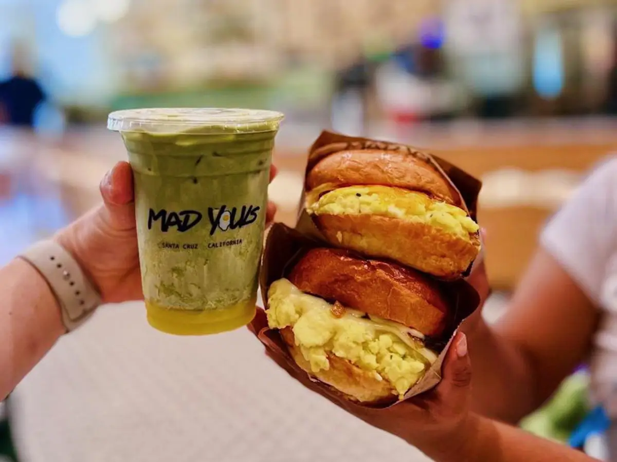 Two hands hold up food items. The hand on the left holds a green beverage in a clear cup labeled "MAD YOUS." The hand on the right holds two egg sandwiches on croissants, showcasing why it's known as the best breakfast in Santa Cruz. The background is blurred, suggesting an indoor setting such as a café or restaurant.