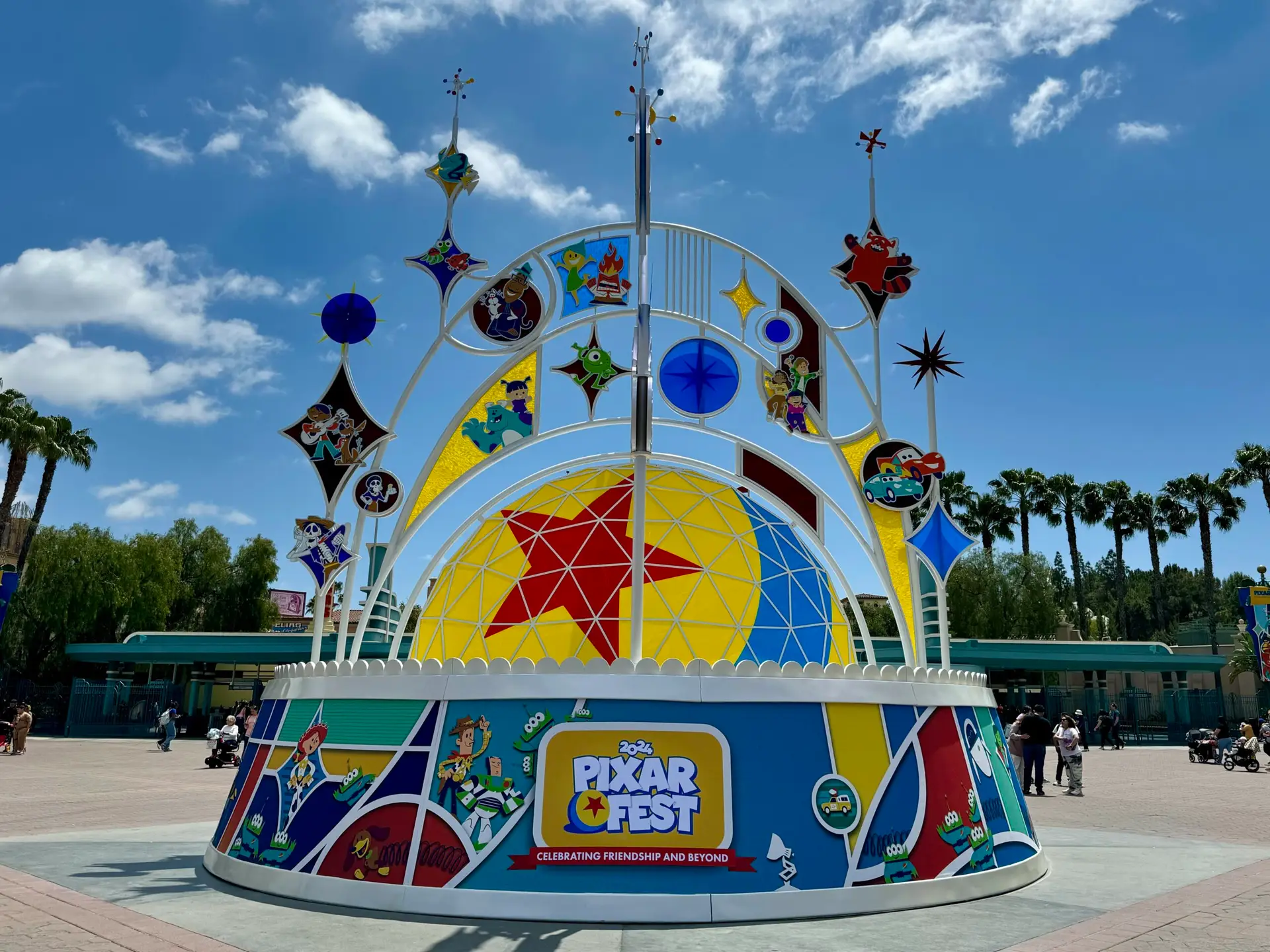 A colorful display at Pixar Fest features various beloved Pixar characters and icons arranged on an arch structure. The background includes a clear sky with some clouds, palm trees, and park greenery. A sign reads, "Pixar Fest: Celebrating Friendship and Beyond.