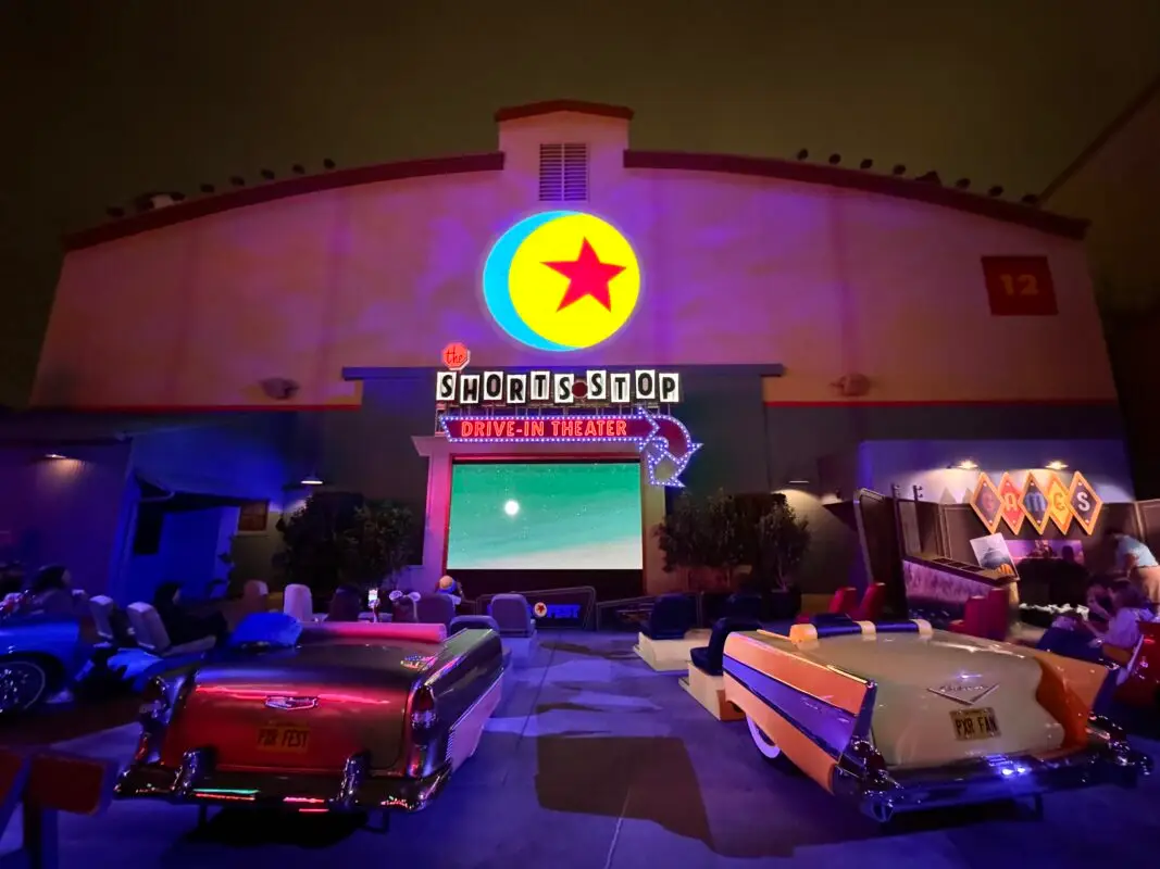 An indoor drive-in theater setup, featuring retro cars facing a large screen. Above the entrance is a brightly lit sign that reads, "Shorts Stop Drive-In Theater," with a neon rocket ship. The ambiance, enhanced by Pixar Fest decorations, is a blend of vintage and futuristic styles.