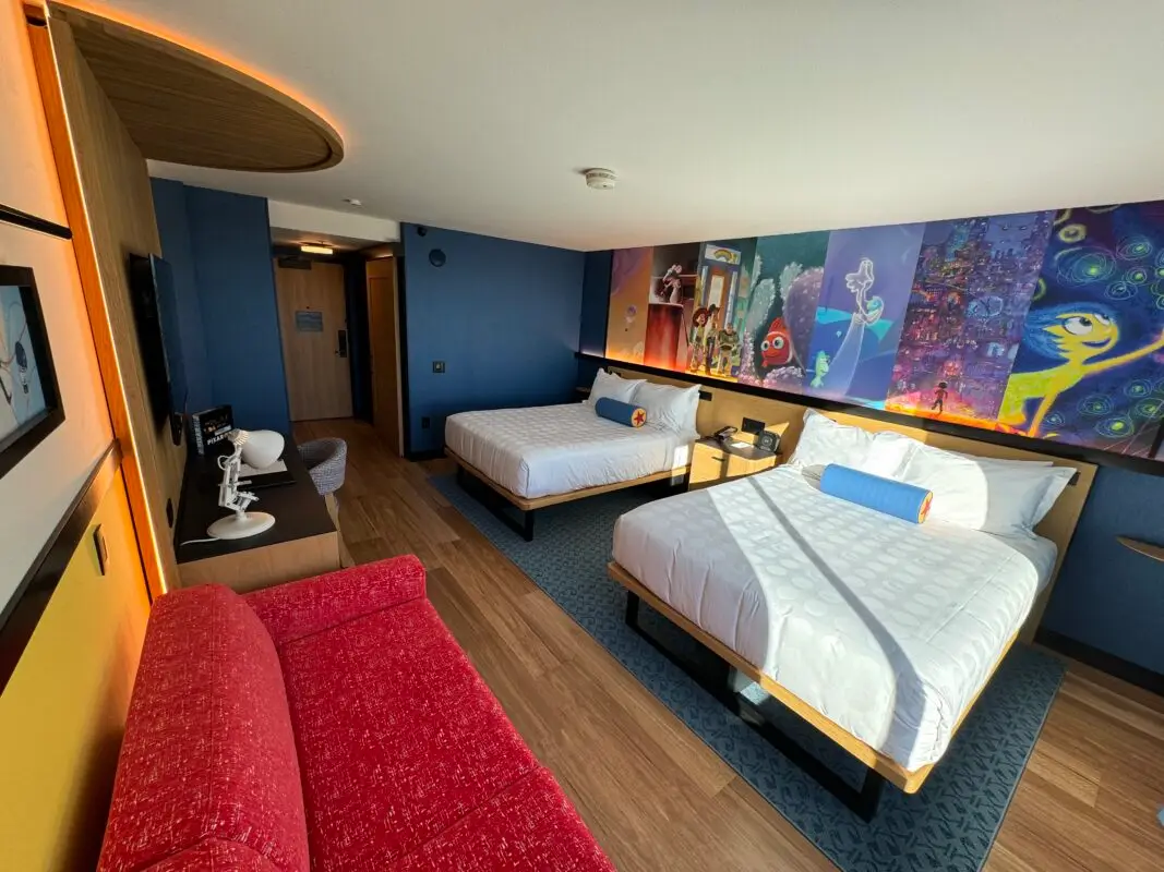A modern hotel room celebrating Pixar Fest features two queen beds with white linens and blue bolster pillows, a red sofa, and a colorful wall mural depicting animated characters. There is a wall-mounted TV, a small desk with two chairs, and wood flooring throughout.