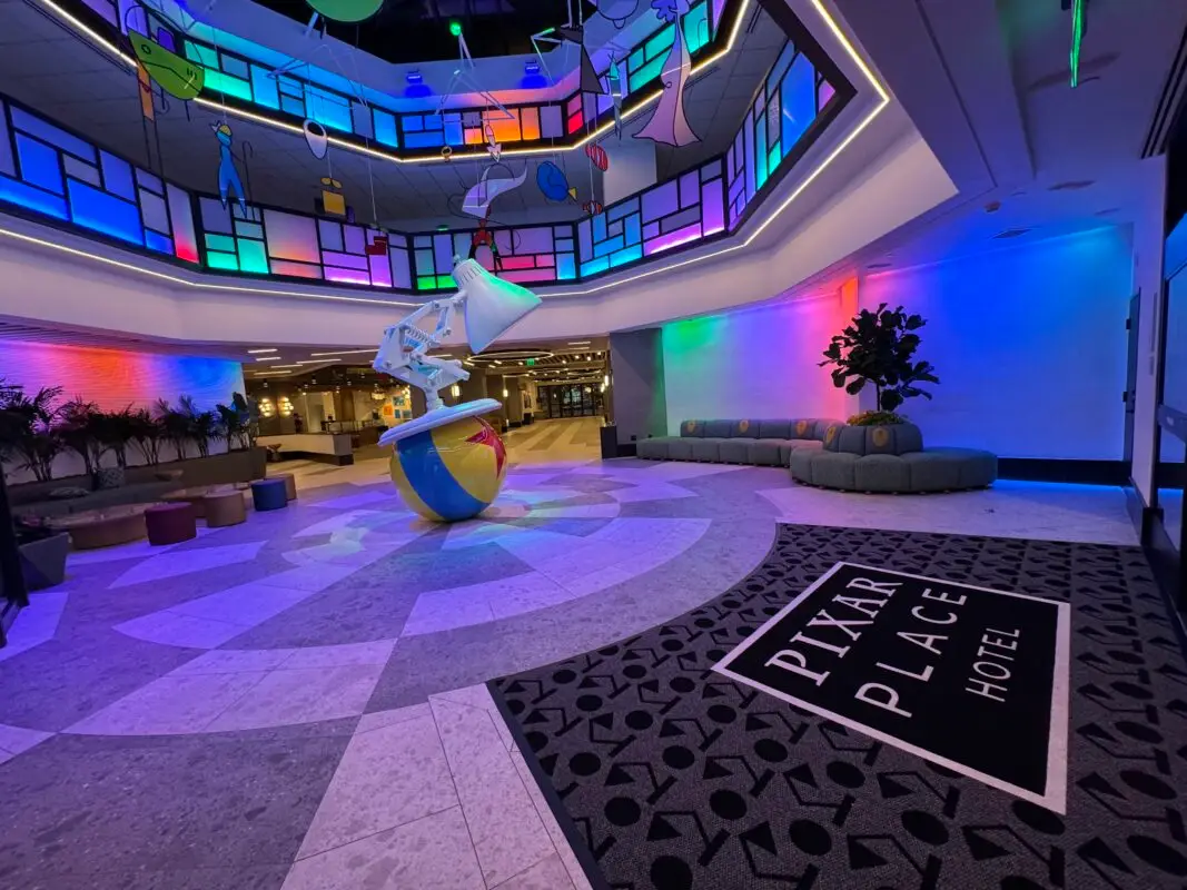 The lobby of Pixar Place Hotel, celebrating Pixar Fest, features a giant Luxo Jr. lamp and ball sculpture under colorful, stained glass-like ceiling lights. Modern seating is arranged around, and a Pixar Place Hotel logo is displayed on a black mat on the floor.