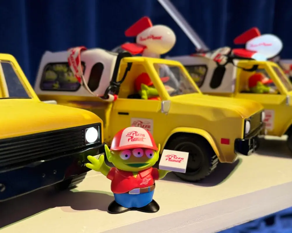 A toy alien wearing a red Pizza Planet cap and shirt is holding a Pizza Planet box while standing in front of a row of toy yellow delivery trucks. The trucks, part of the exclusive Pixar Fest collection, feature detailed designs including a roof-mounted rocket ship and logo decals.