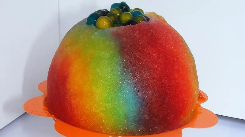 A colorful, dome-shaped gelatin dessert featuring vibrant rainbow hues of red, orange, yellow, green, and blue. The top is adorned with small, circular multicolored candies or fruit pieces. Reminiscent of the best shave ice on the Big Island, it sits beautifully on an orange plate.