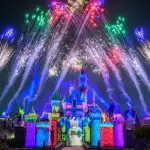 A vibrant nighttime scene at Disney castle during Pixar Fest with fireworks bursting in the sky. The castle is illuminated in various colors, including blue, purple, and pink, while the fireworks display adds more colors like green, orange, and red to the scene.