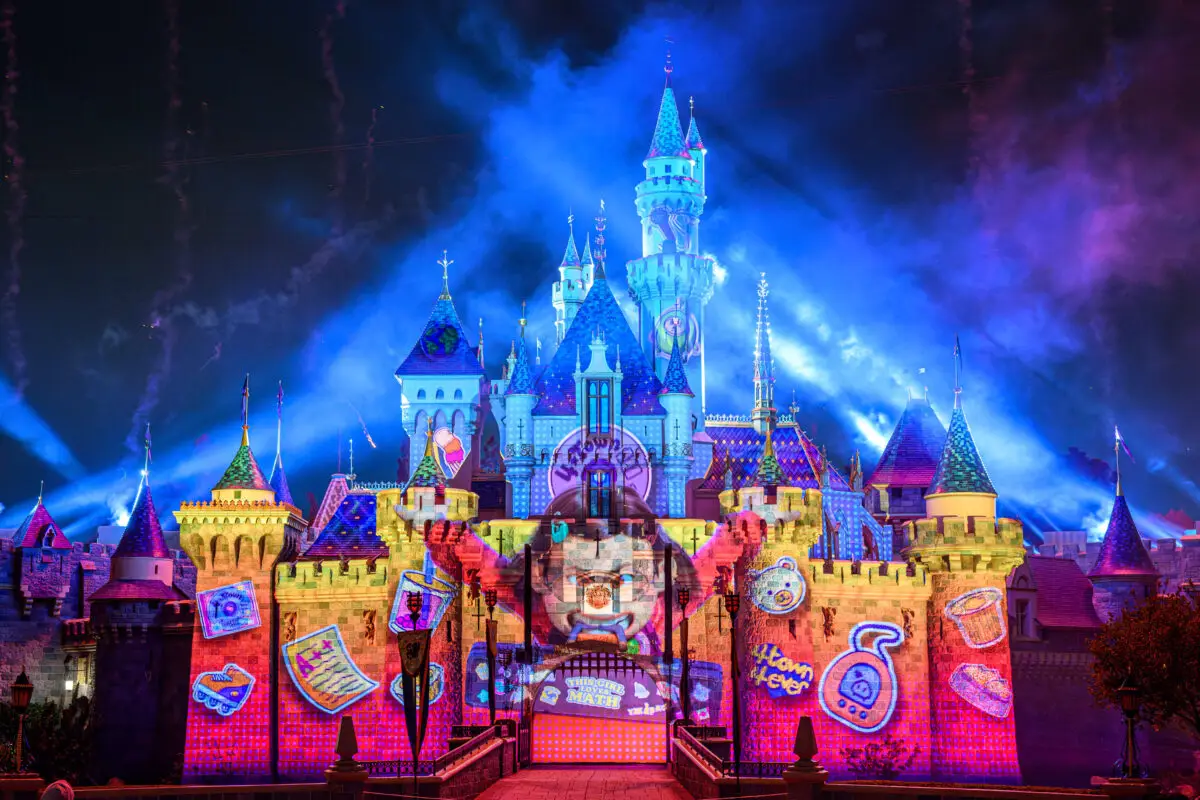 A brightly illuminated castle is colorfully decorated with vibrant projections, featuring various whimsical designs including cartoonish graphs, books, a potion bottle, and a character's face. Blue lights and fireworks enhance the night sky behind the castle during Pixar Fest.