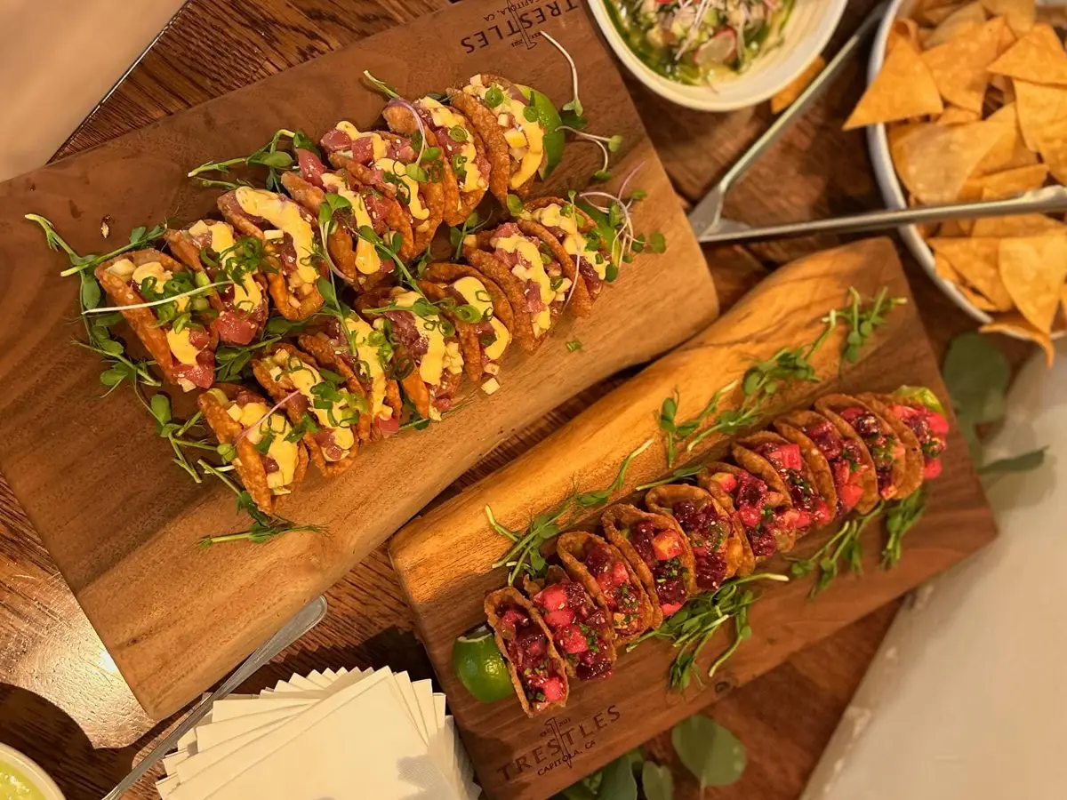 Two wooden boards featuring several mini tacos with various toppings and garnishes, heralding the best dinner in Santa Cruz. The top board has tacos with a cream-colored sauce and microgreens, while the bottom board shows tacos with purple and red toppings. A bowl of chips and dip waits in the background.