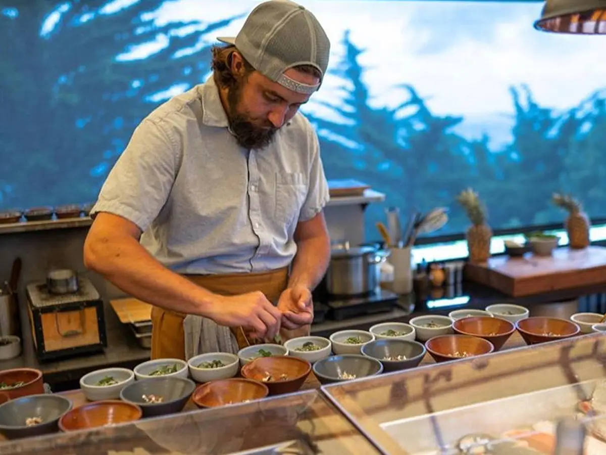 A person wearing a grey cap, light grey shirt, and apron carefully prepares several bowls of food at a kitchen counter. The kitchen has a scenic window view with trees and a cloudy sky in the background. Pineapples and cooking utensils are visible, reminiscent of the best seafood spots in Big Sur.