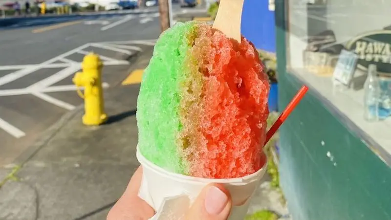 A hand holds the best shave ice on the Big Island in a white cup. The colorful treat is part green and part red with a wooden spoon inserted. In the background, a yellow fire hydrant stands on the sidewalk amid an urban street scene.