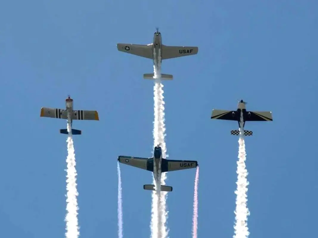 Four military airplanes fly in formation with white smoke trails against a clear blue sky. Two planes in front display "USAF" markings on their wings. Each plane has distinct designs and colors, creating a visually striking scene of aerial precision, perfect for the Memorial Day celebration in the Bay Area.