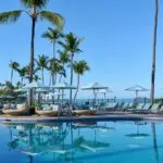 A serene outdoor swimming pool at the Outrigger Kona Resort & Spa is surrounded by lounge chairs and umbrellas under tall palm trees. The clear blue water reflects the cloudless sky and palm trees, exuding a relaxing tropical ambiance.