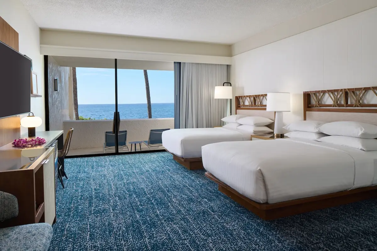 A modern hotel room at the Outrigger Kona Resort & Spa features two large beds, a wall-mounted TV, a desk, and a chair. The room offers a stunning ocean view through its large window and boasts minimalist decor with blue carpet, white walls, and wooden accents.