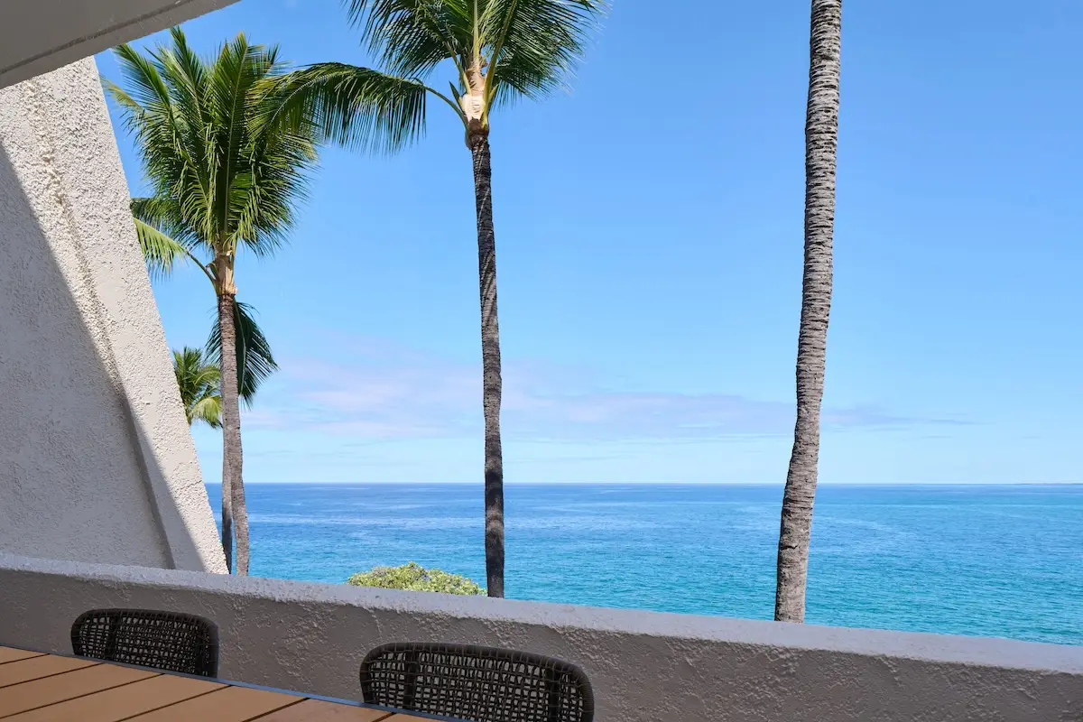 A serene view of a clear blue ocean with a cloudless sky, framed by tall palm trees at the Outrigger Kona Resort & Spa. The scene is viewed from a shaded patio area with wicker chairs and a wooden table in the foreground. The atmosphere is calm and tropical.