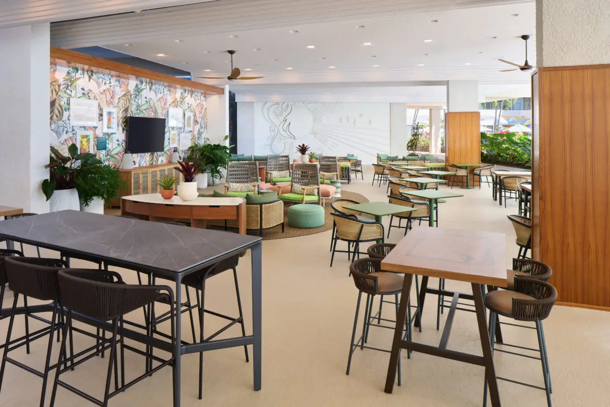 A modern, open-concept café at the Outrigger Kona Resort & Spa offers various seating options like high tables with bar stools, round tables with cushioned stools, and a central area with comfortable chairs. The walls feature vibrant artwork and plants add greenery to the bright, airy space.