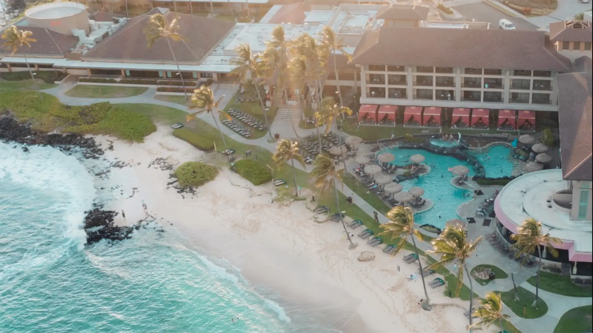 Aerial view of one of the best oceanfront hotels Kauai has to offer, featuring white sand, turquoise water, palm trees, and a large swimming pool surrounded by lounge chairs. Multiple buildings with red canopies and lush green lawns. Guests are relaxing by the pool and beach.