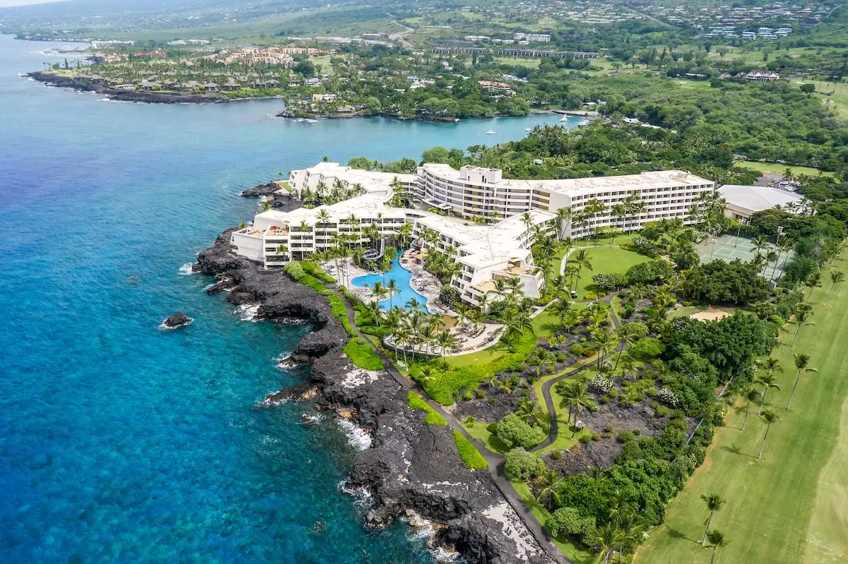 Aerial view of a coastal resort surrounded by lush greenery and clear turquoise waters. The Outrigger Kona Resort and Spa features multiple white buildings, a large swimming pool, and is situated on rocky terrain. Windy roads and a golf course stretch nearby under a partly cloudy sky.