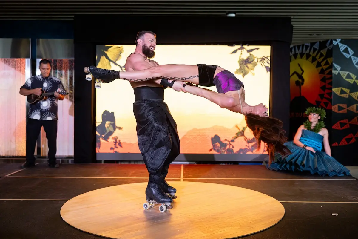 Two performers on roller skates execute a daring lift on a circular wooden platform, with the man holding the woman horizontally as she extends her body. In the background, musicians and a dancer enhance the vibrant stage atmosphere, reminiscent of an ‘Auana performance in Cirque du Soleil Hawaii.