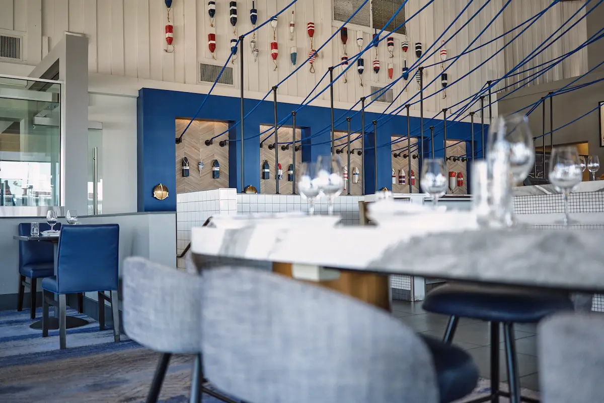 A modern, nautical-themed restaurant interior featuring blue and white decor is perfect for Father’s Day Brunches in the San Francisco Bay Area. Tables are set with wine glasses and napkins, and the ceiling is adorned with colorful float decorations. Blue upholstered chairs and white tiled partitions complement the maritime ambiance.