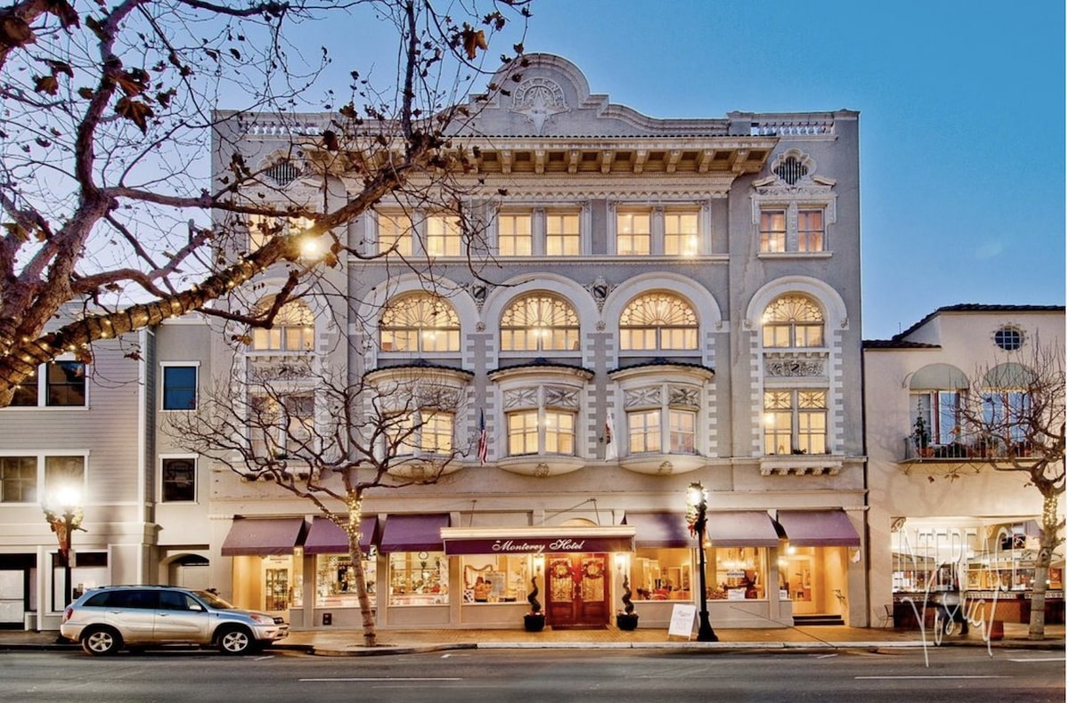 A historic white building with ornate architectural details and arched windows, glowing warmly from interior lights at dusk. The ground floor hosts the "Monterey Hotel," renowned among value hotels, featuring a maroon awning. Leafless trees and parked cars line the street in front.