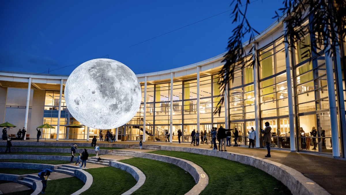 A large artificial moon installation is displayed outside a modern, curved glass building at twilight during one of the Best Annual Events in Napa Valley. People gather around, admiring the art piece. The foreground features tiered concrete seating and grassy areas, and a few children are playing.