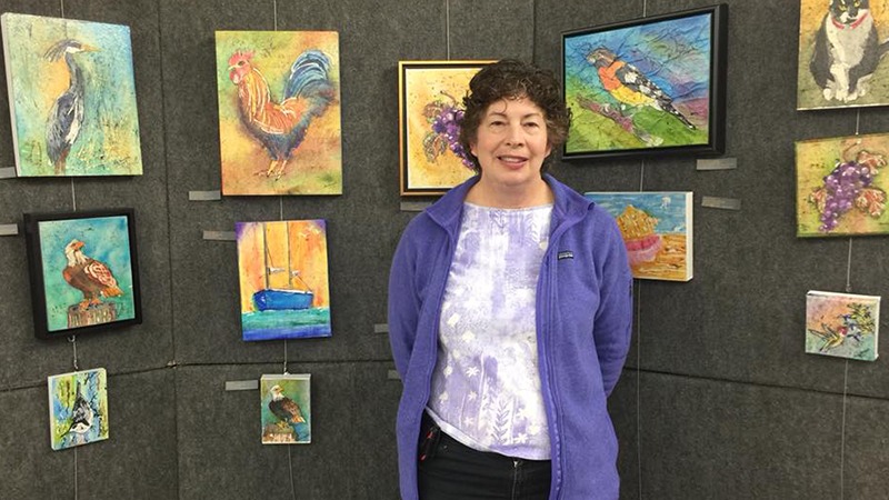 A woman stands in front of an art display featuring various colorful paintings, including birds, a rooster, a boat, and a dog. Wearing a purple jacket and smiling at the camera, she enjoys one of Napa Valley's best annual events. The artwork is hung on a gray wall.