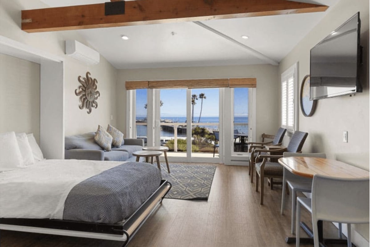 A bright, modern bedroom at Value Hotels Santa Cruz features a bed, sofa, small table, and TV. Large sliding glass doors provide a view of the ocean, palm trees, and patio. The room showcases wood ceiling beams and minimalistic decor with light walls and wooden flooring.