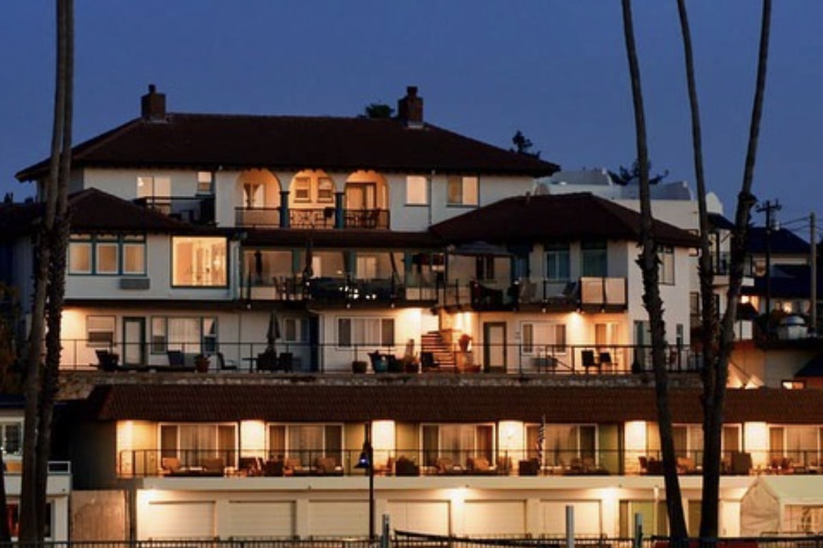 A multi-story building stands illuminated against the evening sky. The structure, part of Value Hotels Santa Cruz, features numerous balconies and large windows, with lights glowing warmly from within. Tall palm trees frame the sides of the building, adding to the serene and picturesque setting.