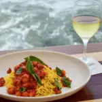 A plate of spaghetti with tomato sauce and basil is set on a wooden table by the sea. A glass of white wine rests next to the plate, which is placed on a white coaster. The background shows waves crashing against the shore—truly, the best lunch Monterey Peninsula has to offer.
