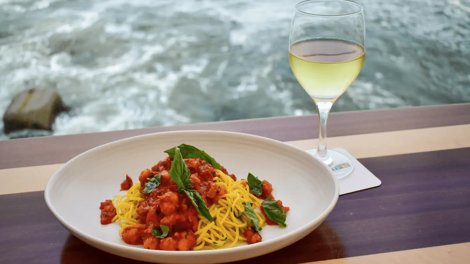A plate of spaghetti with tomato sauce and basil is set on a wooden table by the sea. A glass of white wine rests next to the plate, which is placed on a white coaster. The background shows waves crashing against the shore—truly, the best lunch Monterey Peninsula has to offer.