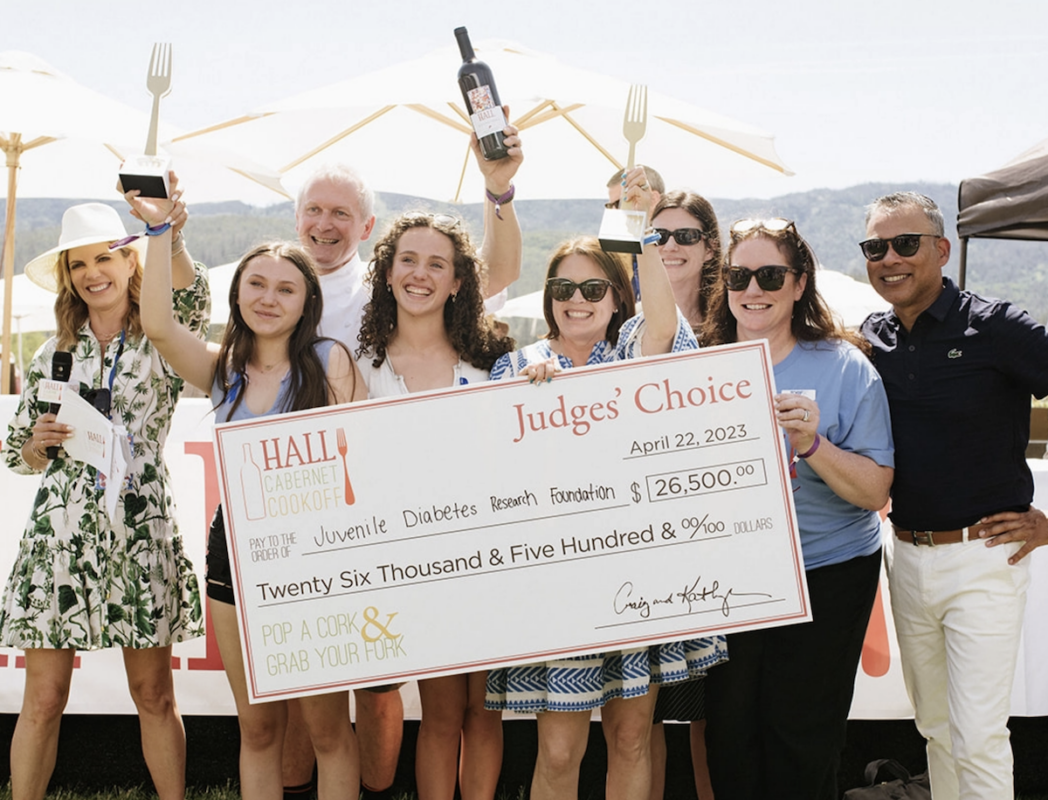 A group of people is holding a large check for $26,500 awarded to the Juvenile Diabetes Research Foundation. They are smiling and standing outdoors at one of Napa Valley's best annual events, the Hall Cabernet Cookoff, on April 22, 2023. One person is holding a wine bottle and microphone.