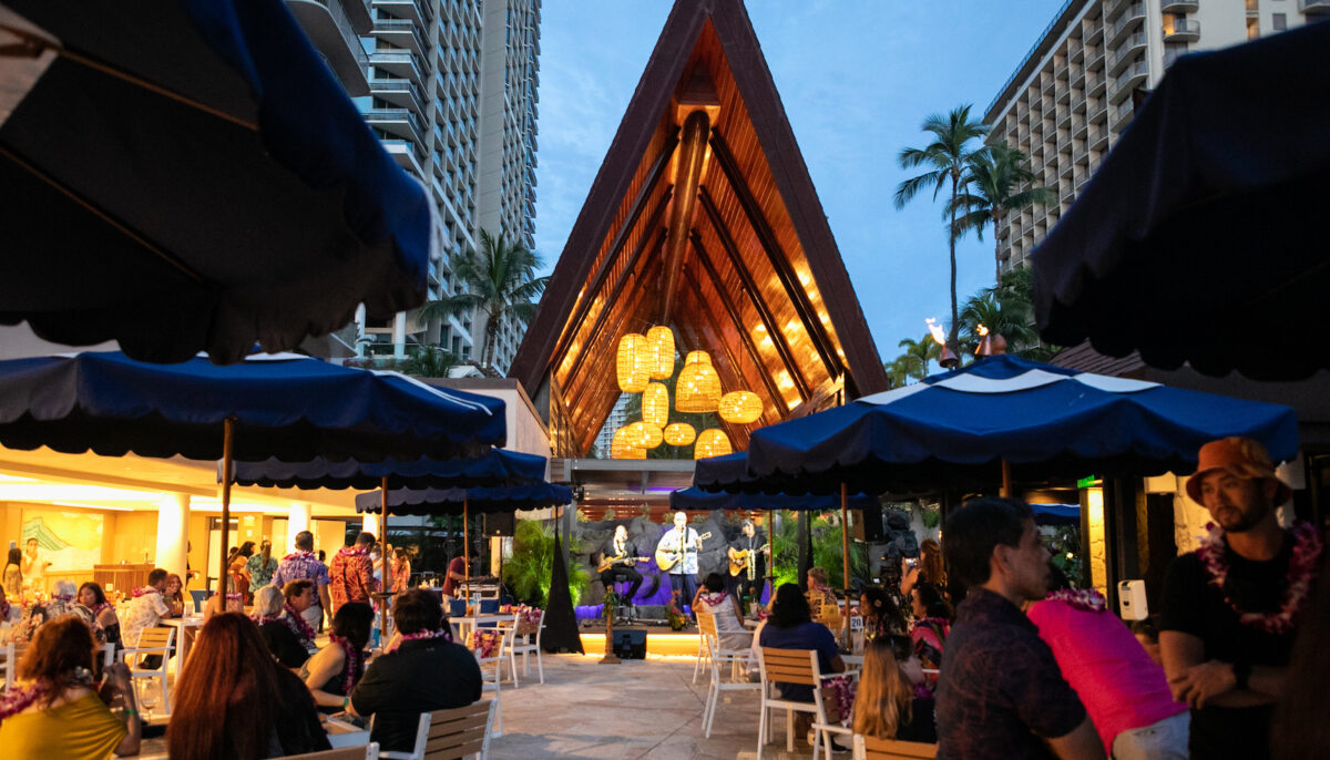 An outdoor evening event at a resort features a live band playing on stage beneath a large wooden A-frame structure. Guests are seated at tables under blue umbrellas, enjoying food and drinks. Palm trees and tall buildings surround the area, creating a tropical atmosphere.