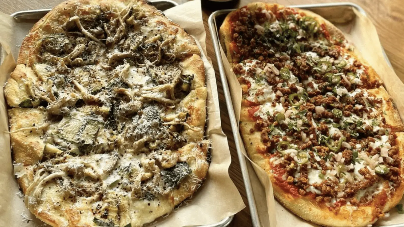 Two rectangular pizzas on metal trays. The left pizza has a white sauce base, mushrooms, shredded cheese, and herbs. The right pizza has a tomato sauce base, ground meat, chopped onions, green peppers, and melted cheese. Both pizzas appear freshly baked—perfect for the best lunch on the Monterey Peninsula.