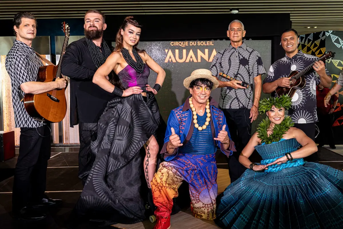 A group of performers from the Hawaiian Cirque du Soleil show, ‘Auana, stands on a stage. They wear colorful, elaborate costumes. Instruments can be seen with a few members, while others pose and smile. A backdrop with the show's title is visible.