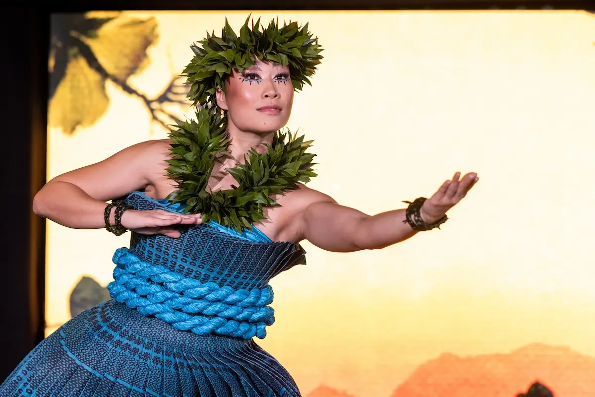 A person wearing a traditional Hawaiian outfit, adorned with a leafy crown and necklace, performs a graceful ‘Auana hula dance. The background features warm shades of yellow and orange, suggesting a sunset or sunrise. The dancer's expression is focused and elegant, reminiscent of Cirque du Soleil's captivating performances.