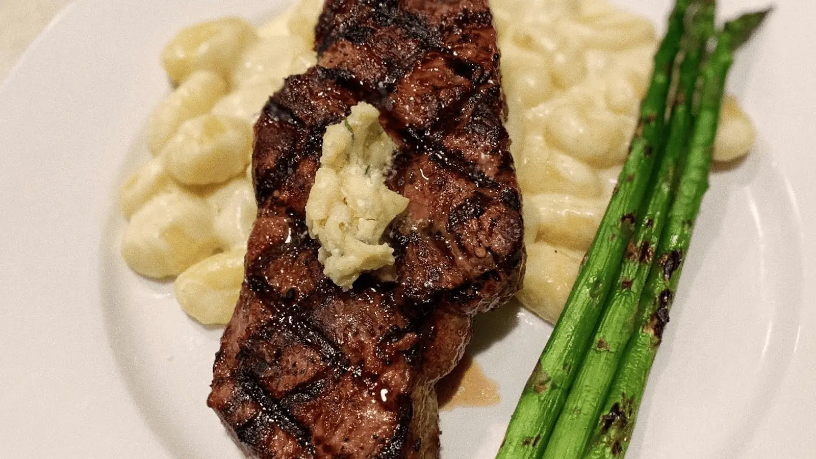 A grilled steak with crisscross grill marks, topped with a dollop of butter, is served alongside creamy gnocchi and steamed asparagus on a white plate—the best lunch you'll find on the Monterey Peninsula.